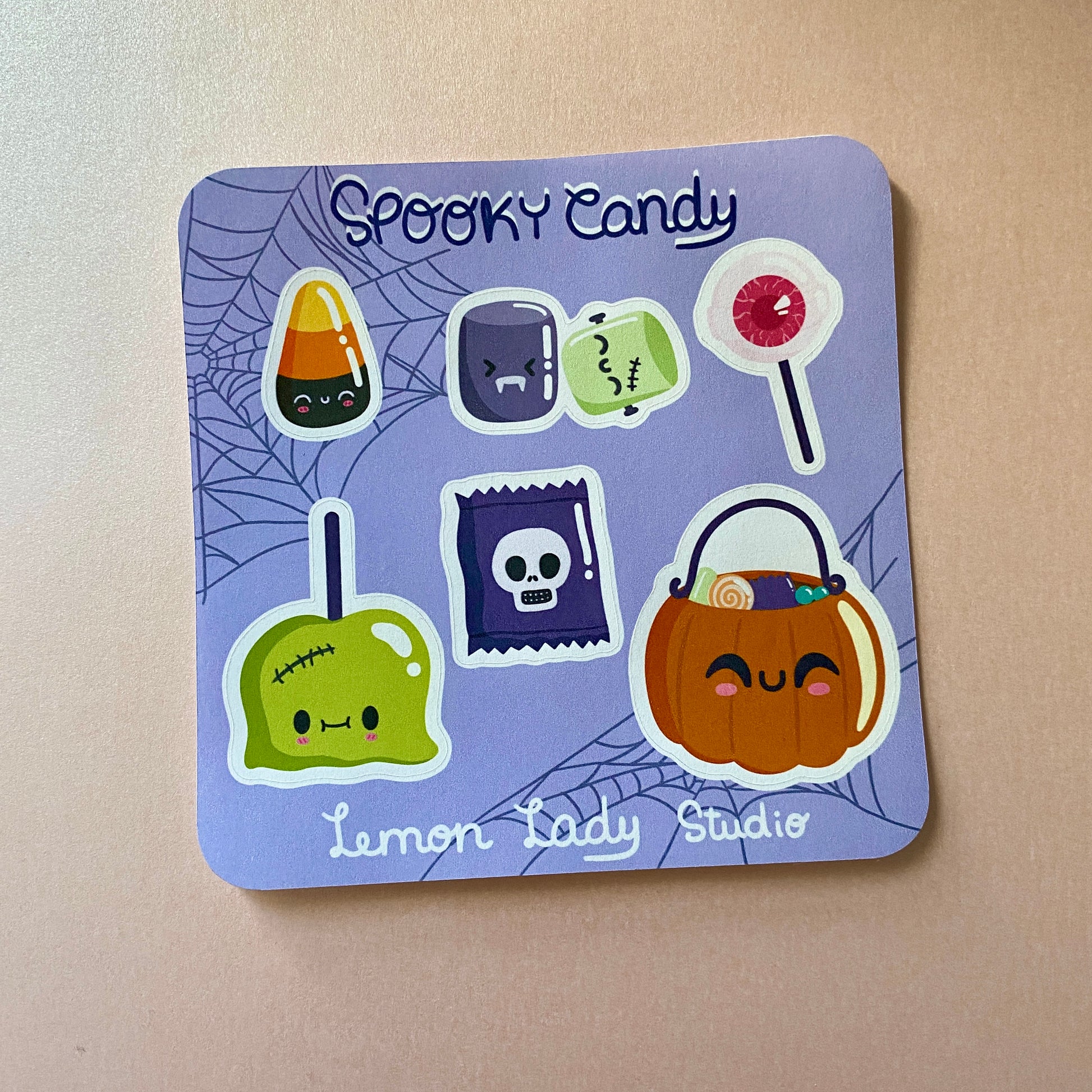 A square Halloween-themed sticker sheet titled "Spooky Candy" on a light purple background with a spiderweb design. The sheet includes six cute and spooky candy designs, such as a candy corn with a smiling face, a Frankenstein-themed caramel apple, a purple candy wrapper with a skull, a green square candy with stitches, a red lollipop, and a pumpkin-shaped candy bucket with a happy expression. The stickers are printed on matte paper, and the "Lemon Lady Studio" logo is visible at the bottom.